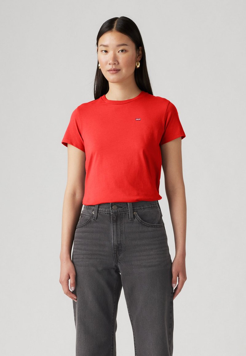 Camiseta de algodón roja con un clásico cuello redondo y mangas cortas, que presenta un pequeño logo azul en el pecho. Combinada con vaqueros grises de cintura alta.