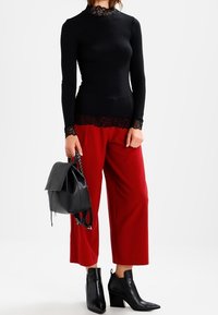 Kvinde iført en langærmet overdel med sort blondedetalje, røde vide bukser, sorte hællet ankelstøvler, mens hun holder en sort læder rygsæk.