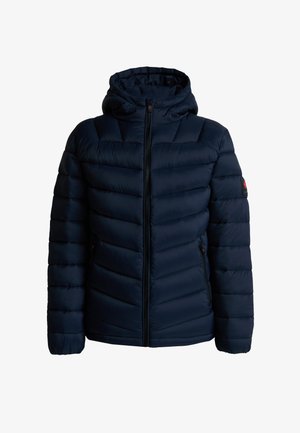 Junge Person mit geflochtenem Haar, die eine marineblaue wattierte Kapuzenjacke, schwarze Jogginghosen und schwarze Sneaker trägt, steht vor einem schlichten hellen Hintergrund.
