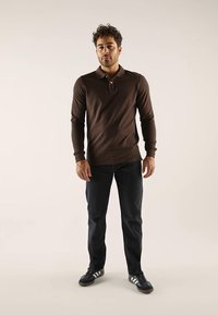Polo-shirt marron à manches longues avec col et trois boutons, associé à un jean bleu foncé et des chaussures de sport noires avec des rayures blanches.