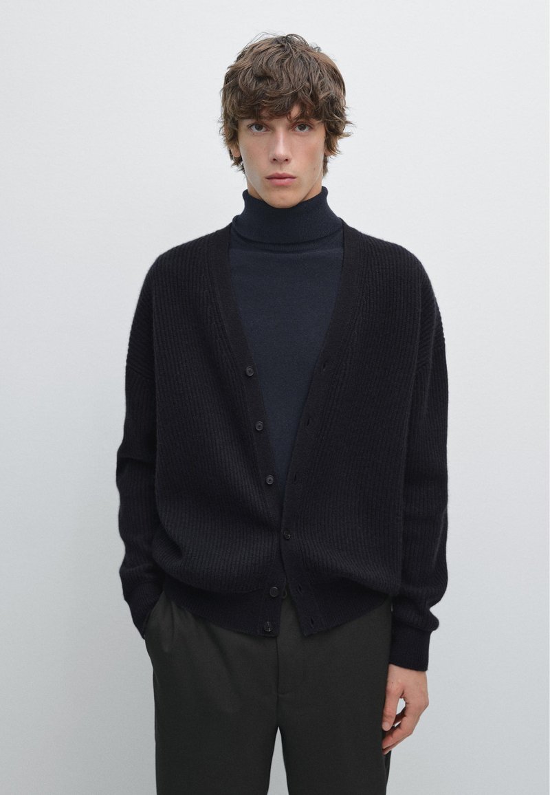 Cardigan nero a coste con scollatura a V e chiusura con bottoni, indossato sopra un dolcevita scuro, con maniche lunghe e vestibilità comoda.