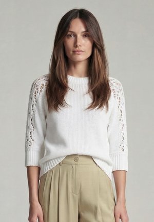 Frau mit langen braunen Haaren, die einen weißen Strickpullover mit Spitzenärmeln und beigen Hosen trägt, steht vor einem schlichten grauen Hintergrund.