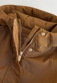 Cuello de chaqueta marrón con cremallera parcialmente abierta, botones de presión metálicos y etiqueta de tela interior visible.