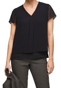 Blusa nera con maniche corte e trasparenti e scollatura a V. Presenta plissettature orizzontali sul collo e un design a strati. Abbinata a pantaloni a fantasia.