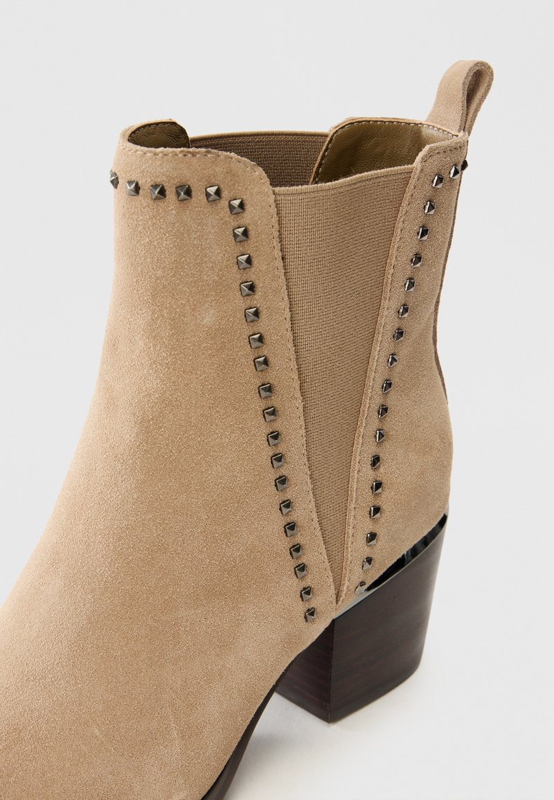 Beżowe zamszowe botki za kostkę z elastycznymi bokami, nakrapianymi akcentami wzdłuż szwów i okrągłym noskiem. Posiadają blokowy obcas dla stabilności.