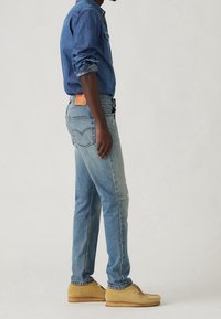 Světle modré džíny z denimu s přímým střihem a vybledlými oblastmi, doplněné o košili z denimu na knoflíky. Obuv z béžové semiše.