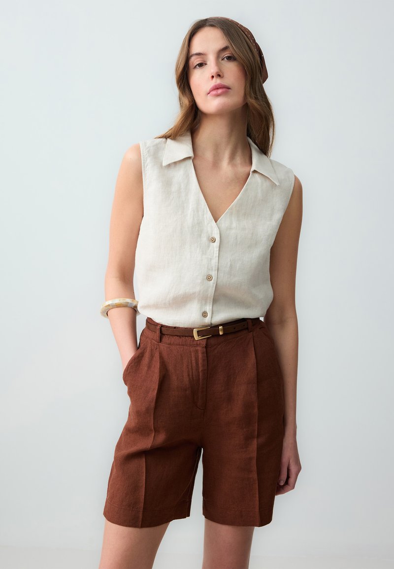Top senza maniche in lino color crema con scollo a V e bottoni, abbinato a pantaloni corti in lino marroni con vita cinturata e pieghe.