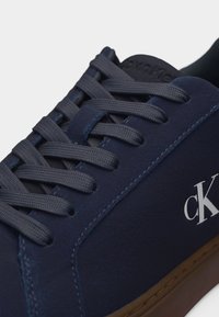Baskets bleu marine avec lacets assortis, semelle marron et logo blanc "ck" sur le côté.