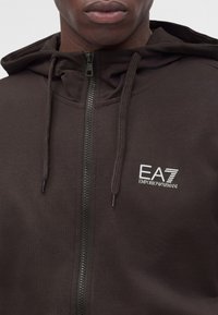 Κοντινό πλάνο ενός ατόμου που φοράει μια σκούρα καφέ φούτερ με φερμουάρ με το λογότυπο "EA7 Emporio Armani" στο αριστερό στήθος και κορδόνια στη κουκούλα.