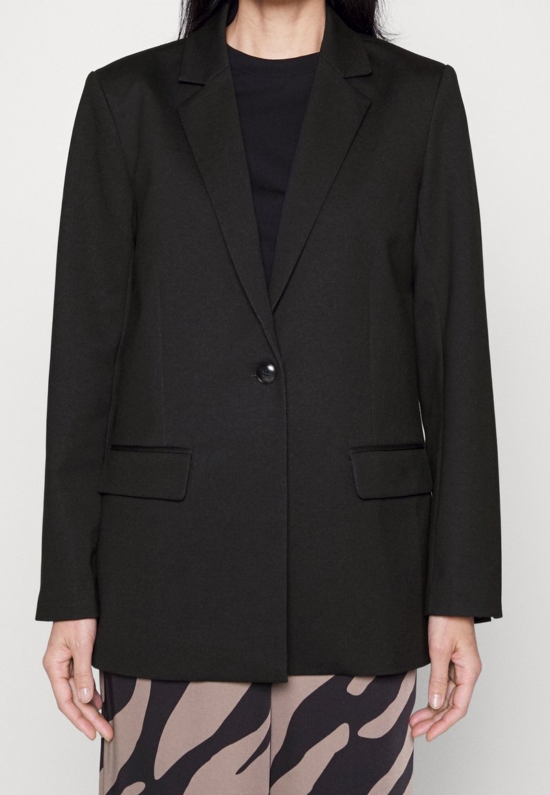 Personne portant un blazer noir à un bouton sur un haut noir et un pantalon beige et noir à motif abstrait, debout devant un fond uni.