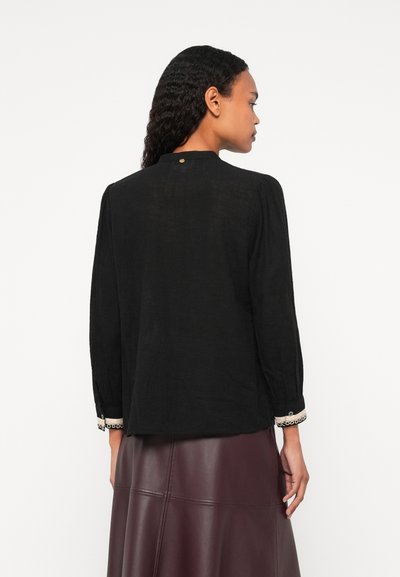 Čierna blúzka s dlhými rukávmi a vysokým golierom, s detailmi gombíkov na zadnej strane a textúrovanou látkou. Rukávy sú ozdobené dekoratívnym lemovaním.