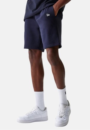 Persona con pantalones cortos azul marino, calcetines blancos hasta la mitad de la pantorrilla y zapatillas blancas, de pie con las manos en los bolsillos contra un fondo liso.