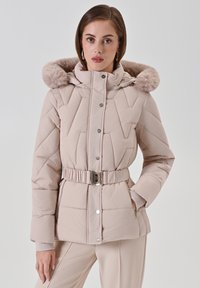 Vrouw in een beige pufferjack met een riem en een bontkraag, staand met één hand in de zak tegen een effen achtergrond.
