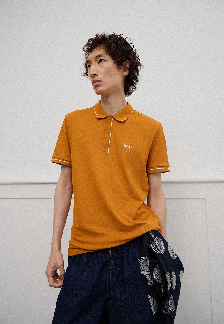 Polo shirt senape con rifinitura bianca al collo e tre bottoni, decorato con un piccolo logo "BOSS" sul petto, abbinato a pantaloni corti blu scuro a motivo.