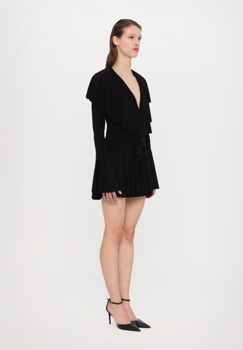 Zwarte jurk met ruches bij de halslijn, lange mouwen en een geplooide rok; gecombineerd met zwarte spitse hakken en een enkelbandje.