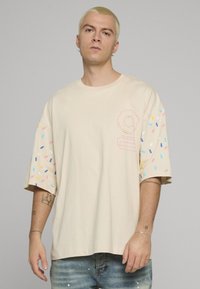 Krémové tričko s krátkými, nadměrně velkými rukávy zdobenými barevnými posypy. Přední strana má jemný růžový motiv. Měkký bavlněný materiál.