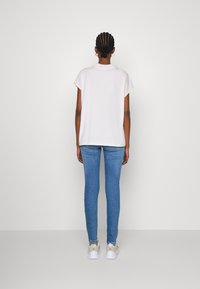 Persona con capelli intrecciati, che indossa una maglietta bianca a maniche corte, jeans skinny blu e sneakers bianche, in piedi di fronte a un muro bianco uniforme.