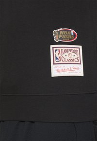 NBA Finals 1996-logotyp ovanför en rektangulär Hardwood Classics Mitchell & Ness-lapp på svart textil.