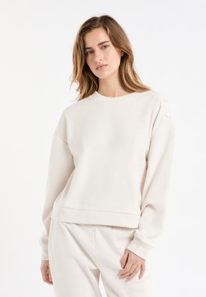 Lichtbeige geribbelde trui met lange mouwen, een ronde hals en decoratieve knopen op de schouder. Zachte textuur, ontspannen pasvorm.