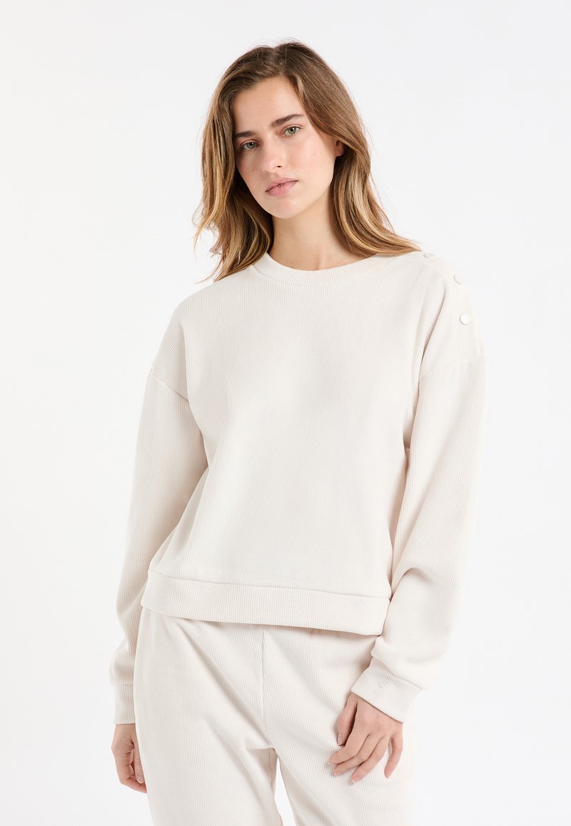 Jasnobeżowa prążkowana bluza z długimi rękawami, okrągłym dekoltem i ozdobnymi guzikami na ramieniu. Miękka tekstura, luźny krój.