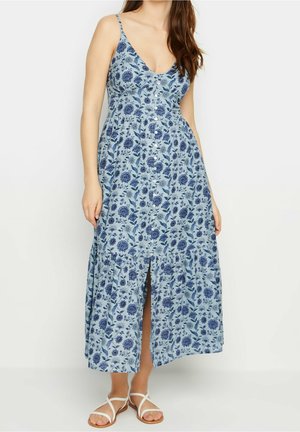 Kvinde iført en lyseblå, ærmeløs maxikjole med mørkeblå blomsterprint, knapper foran og hvide sandaler med stropper.