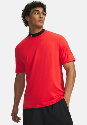 Man met een felrood Under Armour T-shirt en zwarte sportshort, staand met één hand in zijn zak, kijkend opzij.
