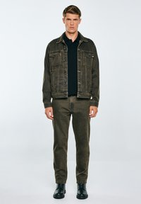 Chaqueta de mezclilla verde oscura con bolsillos frontales, superpuesta a una camiseta polo negra. Unos pantalones verdes oscuros de corte slim y unas botas negras con cordones completan el conjunto.