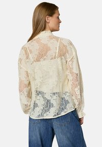 Kvinde med langt hår iført en gennemsigtig cremefarvet blonderbluse over en top og løse blå jeans, vendt sidelæns mod en ensfarvet baggrund.