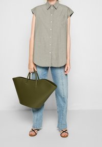 Maglietta grigia senza maniche, jeans blu chiaro, grande borsa tote verde oliva con design strutturato e sandali neri con cinturini.
