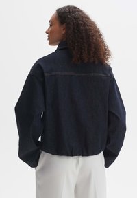 Veste en denim foncé avec une coupe décontractée, des épaules tombantes et un ourlet élastique. Présente des coutures visibles et un design de col classique.
