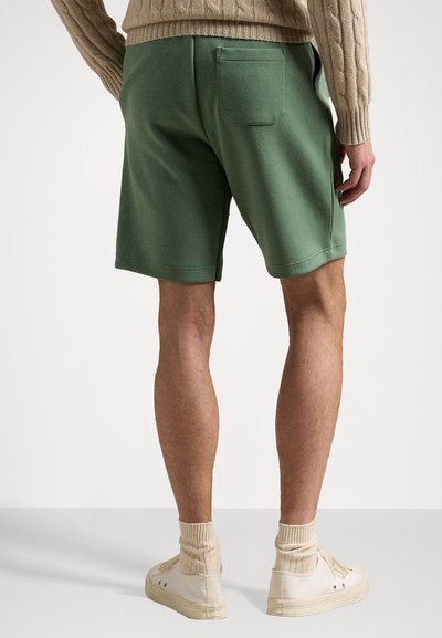 Calções de algodão verdes com um bolso traseiro, combinados com ténis de cano alto creme e meias bege caneladas, exibidos de costas.