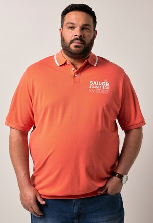 Koralfarvet polo t-shirt med hvid krave og forfriskende tryk; kortærmet og afslappet pasform; iført med blå jeans og et ur.