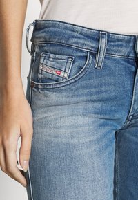 Siniset farkut, joissa on haalistunut pesu, kontrastivärinen ommel ja pieni logo-tagi etutaskussa. Leikkaus on slim fit, mikä korostaa muotoa.