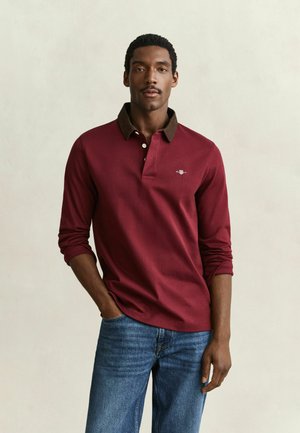 Uomo che indossa una polo bordeaux a manica lunga con colletto marrone e jeans blu, in piedi con una mano in tasca davanti a uno sfondo semplice.