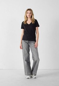 T-shirt nera con scollatura a v abbinata a jeans larghi grigi chiari. Modella che indossa sneakers sportive grigie. Tessuto dalla consistenza liscia e vestibilità casual.