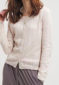Licht beige brei cardigan met een gestructureerd patroon, ronde halslijn en knoopsluiting. Beschikt over lange mouwen en een zachte, ademende stof.