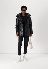 Sort dunjakke med blank finish og stor krave, båret over en sort top, parret med mørke jeans og hvide sneakers. Tilbehør inkluderer solbriller og en lille taske.