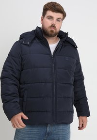 Mørkeblå puffjakke med hætte, der har en frontlynlås, kviltet design og sidelommer. Lavet af et glat, vandafvisende materiale.