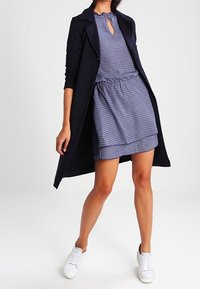 Marineblauer Trenchcoat über einem blauen gemusterten Kleid mit einem Schlüssellochausschnitt, tailliertem Schnitt und weißen Sneakers auf hellem Hintergrund.
