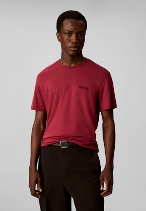 Mand iført en rød T-shirt med korte ærmer og sorte bukser med sort bælte mod en ensfarvet lys grå baggrund.