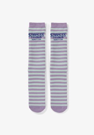 Chaussettes rayées violet et bleu clair avec le logo "Stranger Things Benetton" près du haut, posées à plat côte à côte.