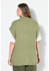 Frau mit langen Haaren, großen Creolen, einer grünen ärmellosen Strickweste über einem weißen T-Shirt und grünen gemusterten Hosen, von hinten gesehen.