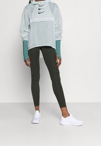 Cazadora ligera color verde menta con tres logotipos negros de Nike, combinada con leggings verde oscuro y zapatillas deportivas blancas. Mangas largas con agujeros para los pulgares.