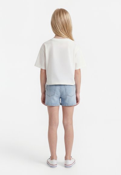 Bērns ar gariem blondiem matiem stāv ar muguru, valkā baltu īsroku krekliņu, gaišas džinsa šortus un baltus sporta apavus uz balta fona.
