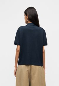 Γυναίκα με μακριά σκούρα μαλλιά, φορώντας μπλε σκούρο πουκάμισο με κοντά μανίκια και μπεζ παντελόνι με φαρδιά πόδια, γυρισμένη από την πλάτη, μπροστά σε απλό ανοιχτό φόντο.