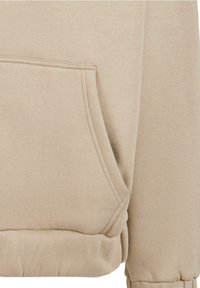 Lys tan hoodie med kængurulomme, ribbenede manchetter og kant, blød tekstur og syninger. Ingen mønstre eller logoer synlige.