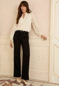 Blusa bianca a pois con maniche a sbuffo, scollo a V, abbinata a jeans neri a vita alta a zampa d'elefante e décolleté crema. Design elegante con dettagli testurizzati.