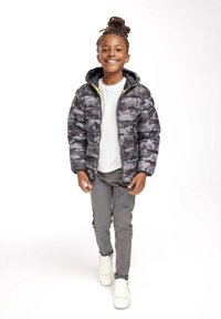 Giacca puffer mimetica grigia con cappuccio, cerniera gialla, indossata sopra una maglietta bianca. Abbinata a pantaloni grigi e sneakers bianche.