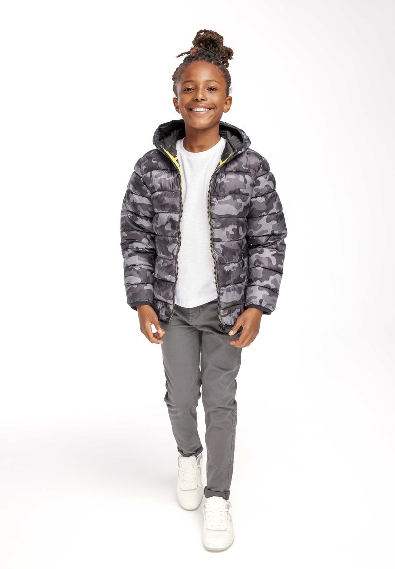 Giacca puffer mimetica grigia con cappuccio, cerniera gialla, indossata sopra una maglietta bianca. Abbinata a pantaloni grigi e sneakers bianche.