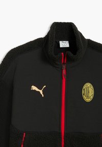 Veste polaire noire avec fermeture éclair rouge, logo Puma doré sur la poitrine gauche et emblème doré AC Milan 1899 sur la poitrine droite.